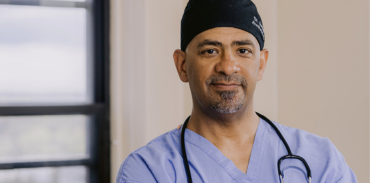 Dr. Colin Audain wearing blue scrubs wearing a stethoscope around his neck and black scrub cap