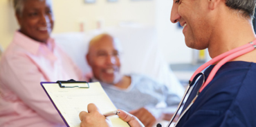 nurse writing notes on a clipboard while a hospitalized man watches with a female companion