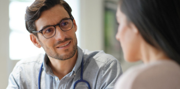 male physician with short brown hair wearing a grey shirt, glasses, and a stethoscope around his neck speaking to a female patient with long brown hair and beige sweater