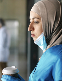 woman physician wearing a hijab and blue scrubs and mask, holding a coffee cup while deep in thought