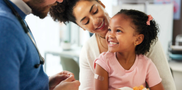 Smiling female child sitting on her mothers lap. Child has a bandage on her upper arm and is looking up at a male physician.
