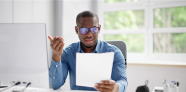 Black man wearing blue shirt and looking at a piece of paper in his hand and looking confused while sitting at a desk with a computer monitor