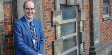 Man wearing blue suit and tie leaning against a brick wall