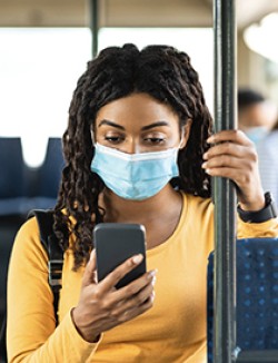 Black woman standing on a bus while looking at her phone and wearing a mask