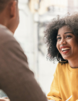 Black woman wearing a yellow sweater smiling while speaking to man wearing a brown sweater