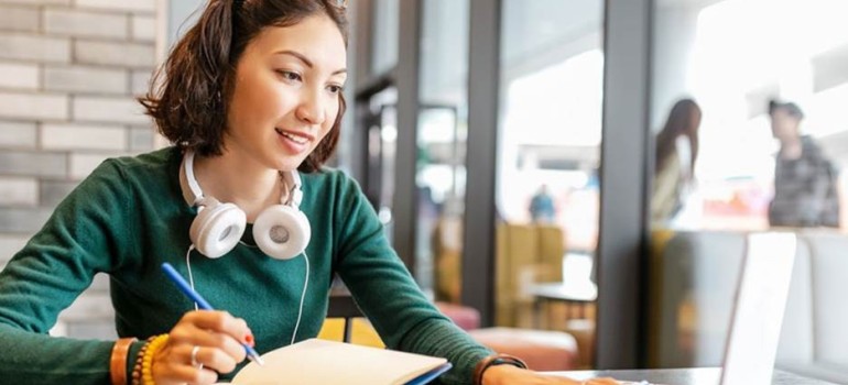 Female student wearing a green sweater, headphones around her neck and glasses on her head. She's taking notes on paper from a laptop computer