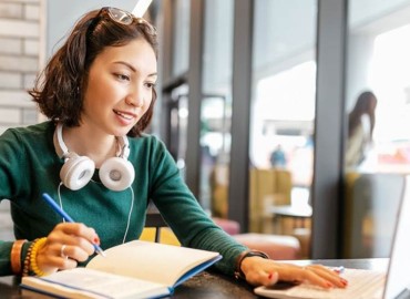 Female student wearing a green sweater, headphones around her neck and glasses on her head. She's taking notes on paper from a laptop computer