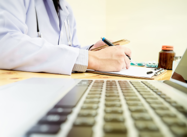physician at desk writing with a laptop keyboard beside them