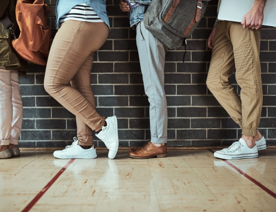 image of various people's legs with different style of pants, waiting in a line behind each other against a brick wall