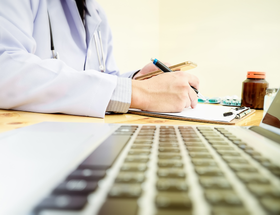 physician at desk writing with a laptop keyboard beside them